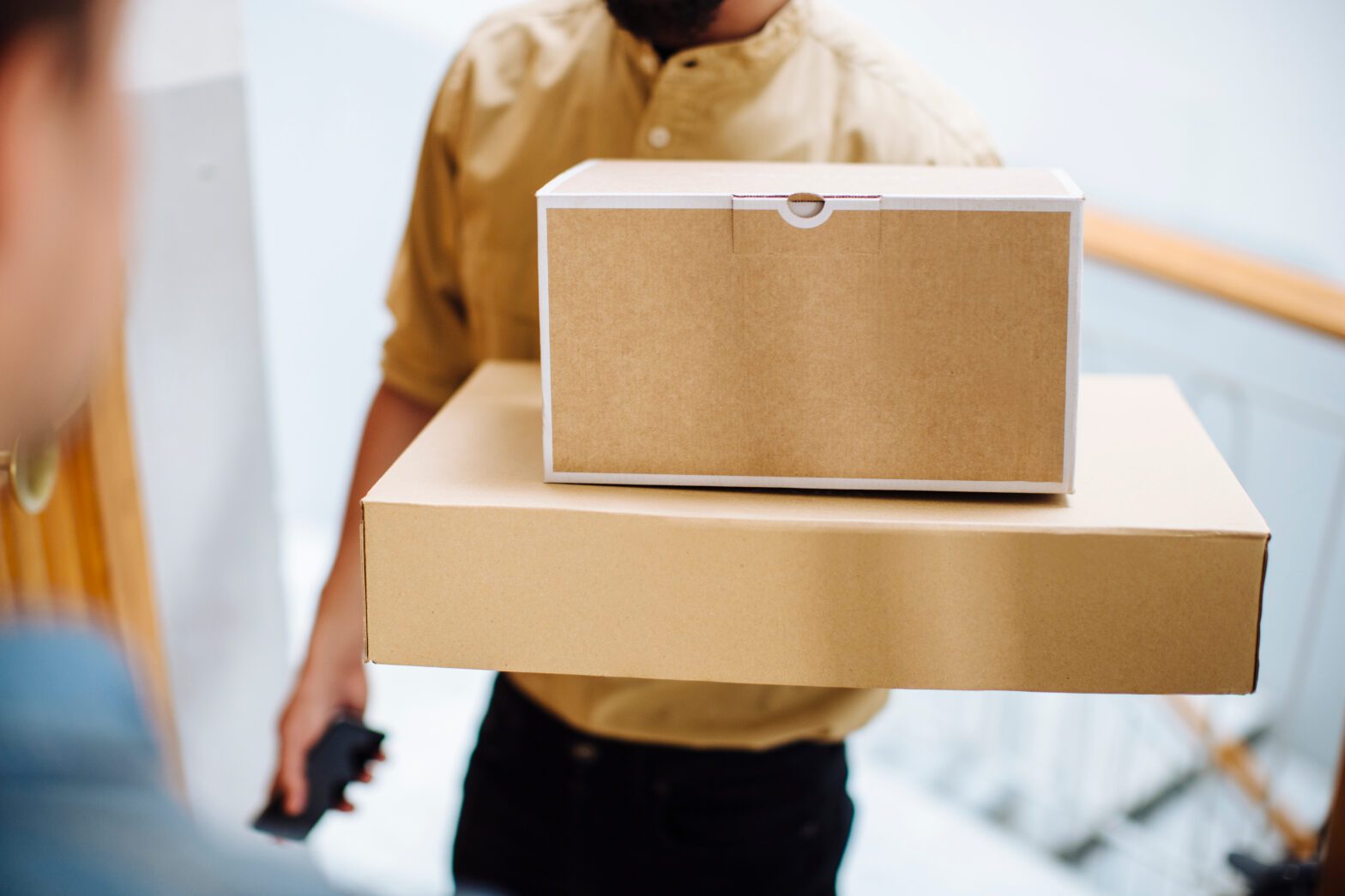 Image of delivery person in a yellow shirt with dark pants handing two boxes stacked on top of one another to the recipient.