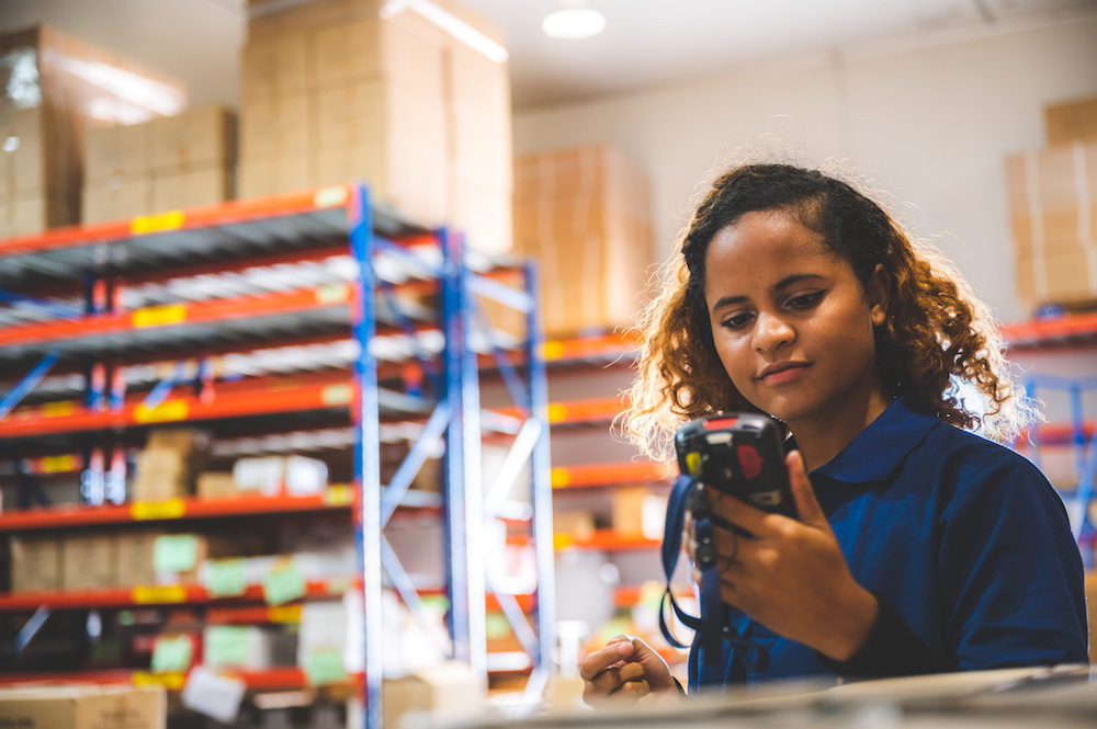 A woman working in a 3PL warehouse, standing in front of racks of products and looking down at a scanner.